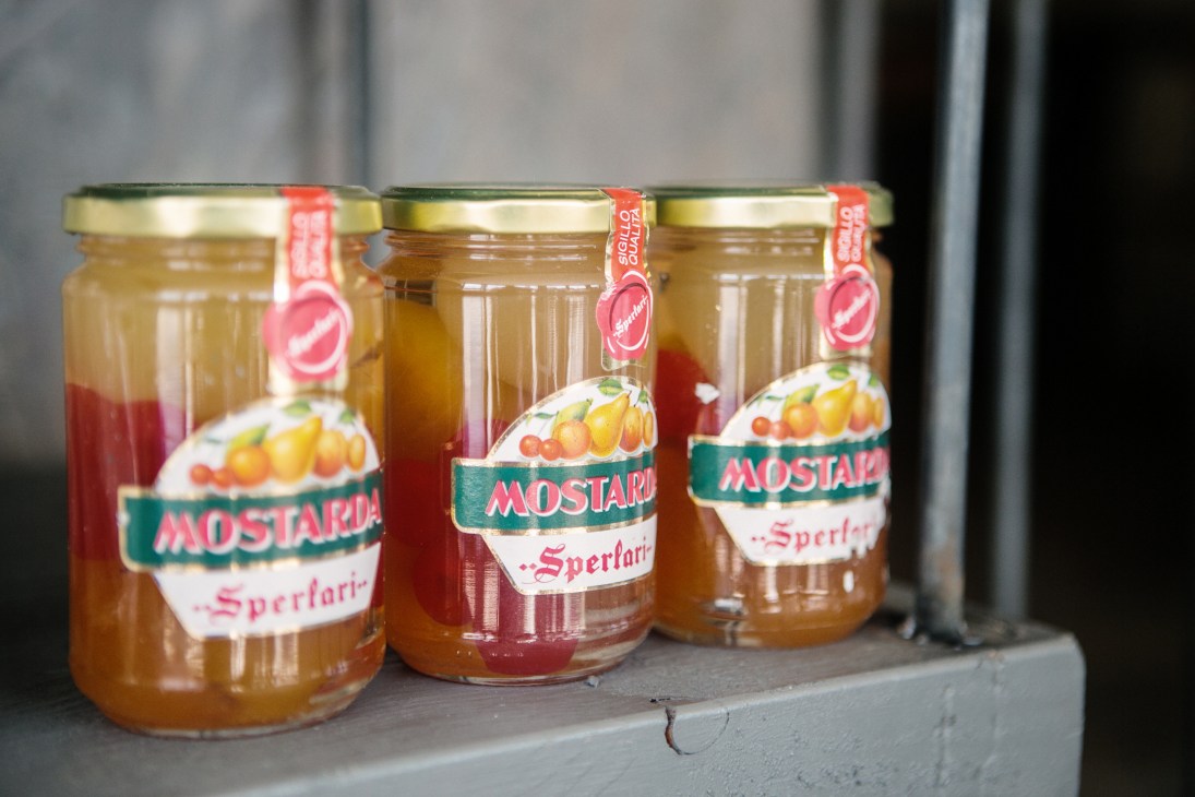 Three jars of Mostarda Sperlari preserves with red ribbon labels on a grey surface.