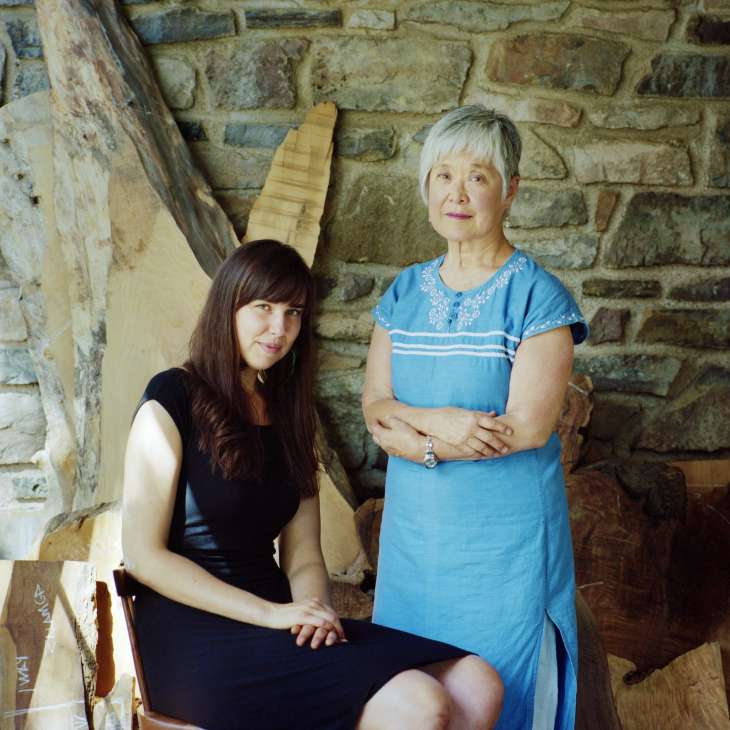 Miriam Carpenter and Mira Nakashima stand together in their studio with a stone wall and wooden furniture pieces behind them.