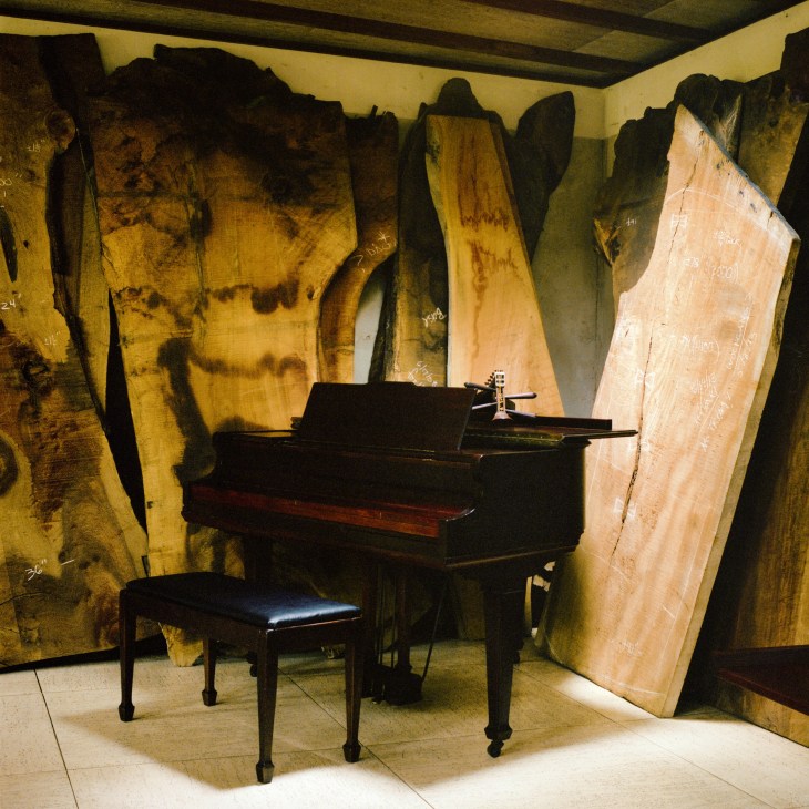 A black grand piano surrounded by large wood slabs with varied grain patterns leaning against walls in a studio space.