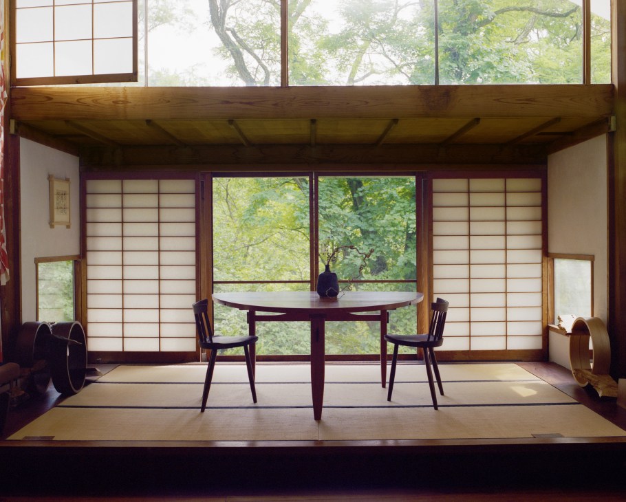 Minimalist dining room with a round dark wood table and two chairs overlooking a forest through large windows.