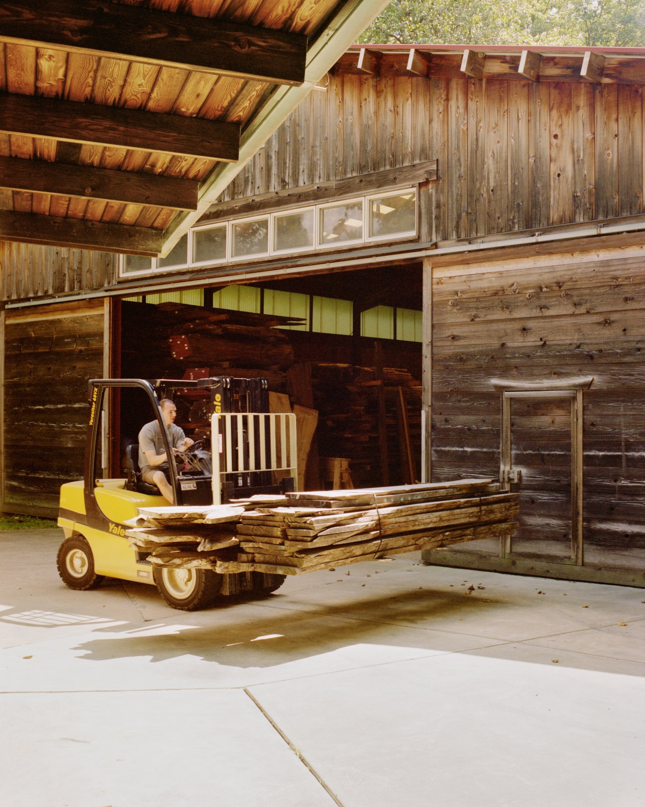 A worker operates a yellow forklift carrying wooden planks outside a wooden workshop building at Nakashima Woodworker.
