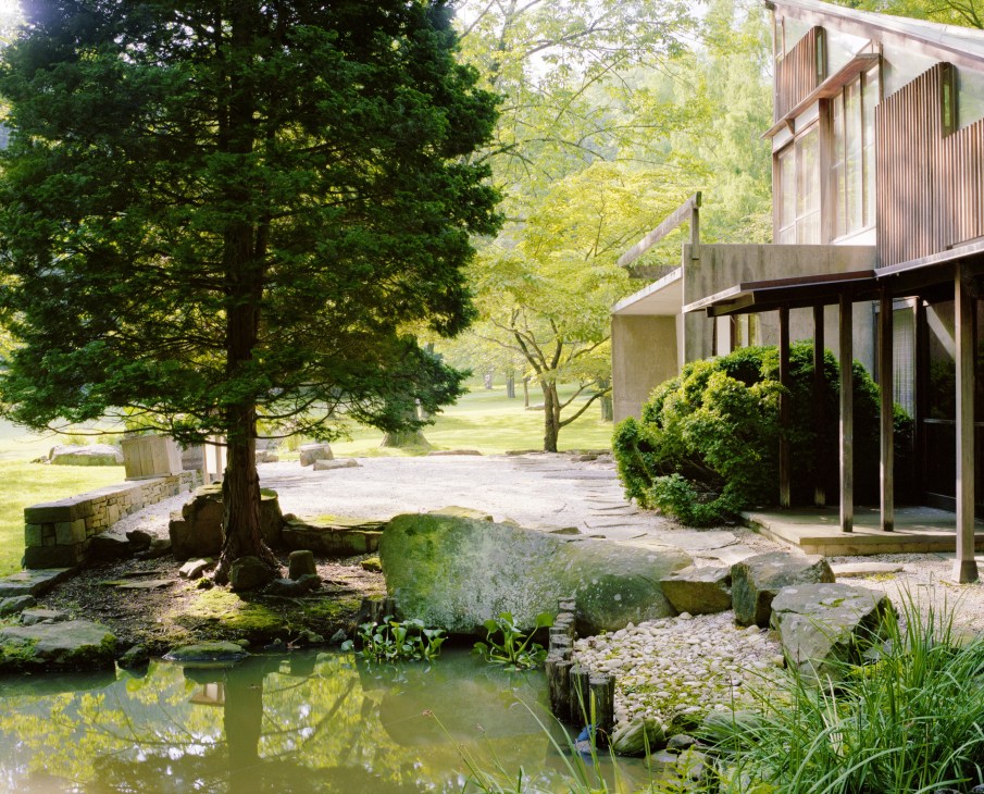 Exterior view of a modernist home with concrete and wood siding set among mature trees and a pond on the Nakashima Woodworker property.