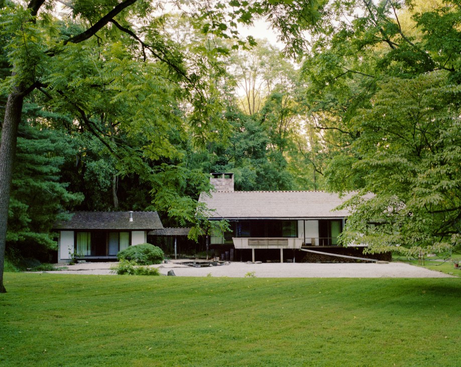 Modern residential home with horizontal lines nestled among mature trees, featuring a chimney and expansive lawn in foreground.