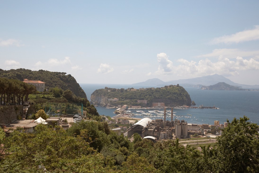 View of Naples Bay with Vesuvius volcano visible across the water and a coastal city sprawl below.