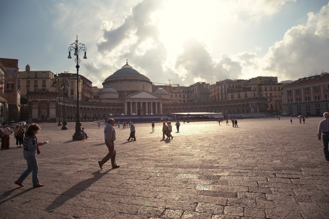 Piazza del Plebiscito in Naples with neoclassical dome and colonnade, people scattered across the square under cloudy sky.