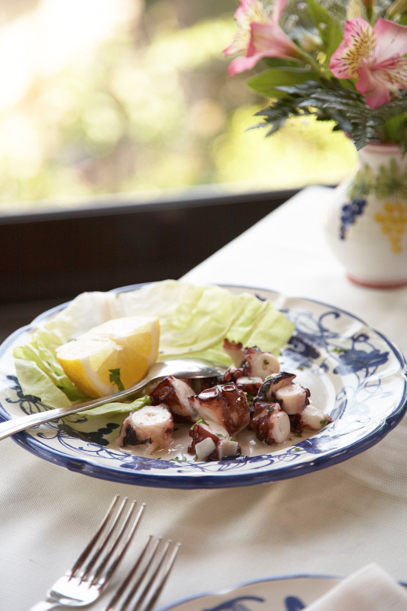 Insalata Polpo dish with octopus, lemon wedge and lettuce on a blue and white plate with flowers in background