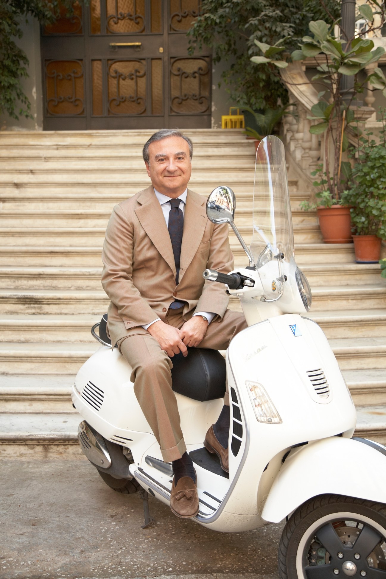 Man in beige suit sitting on a white Vespa scooter in front of a grand doorway.