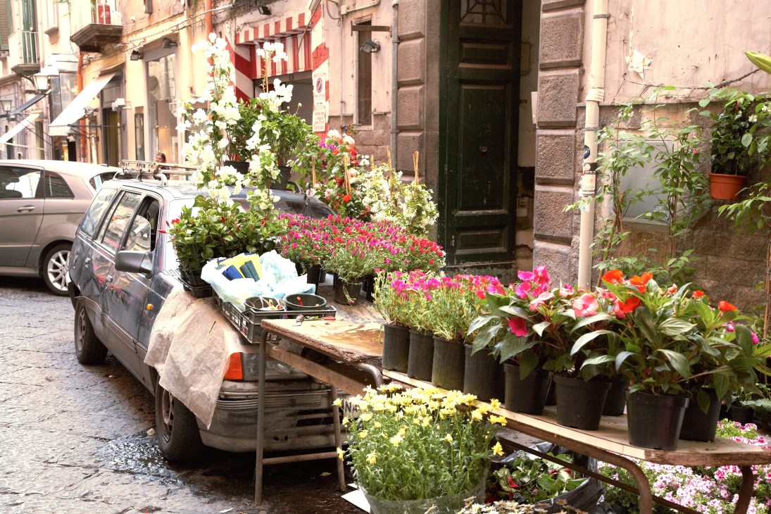 A flower vendor's three-wheeled vehicle laden with potted plants and flowers on a narrow Naples street.
