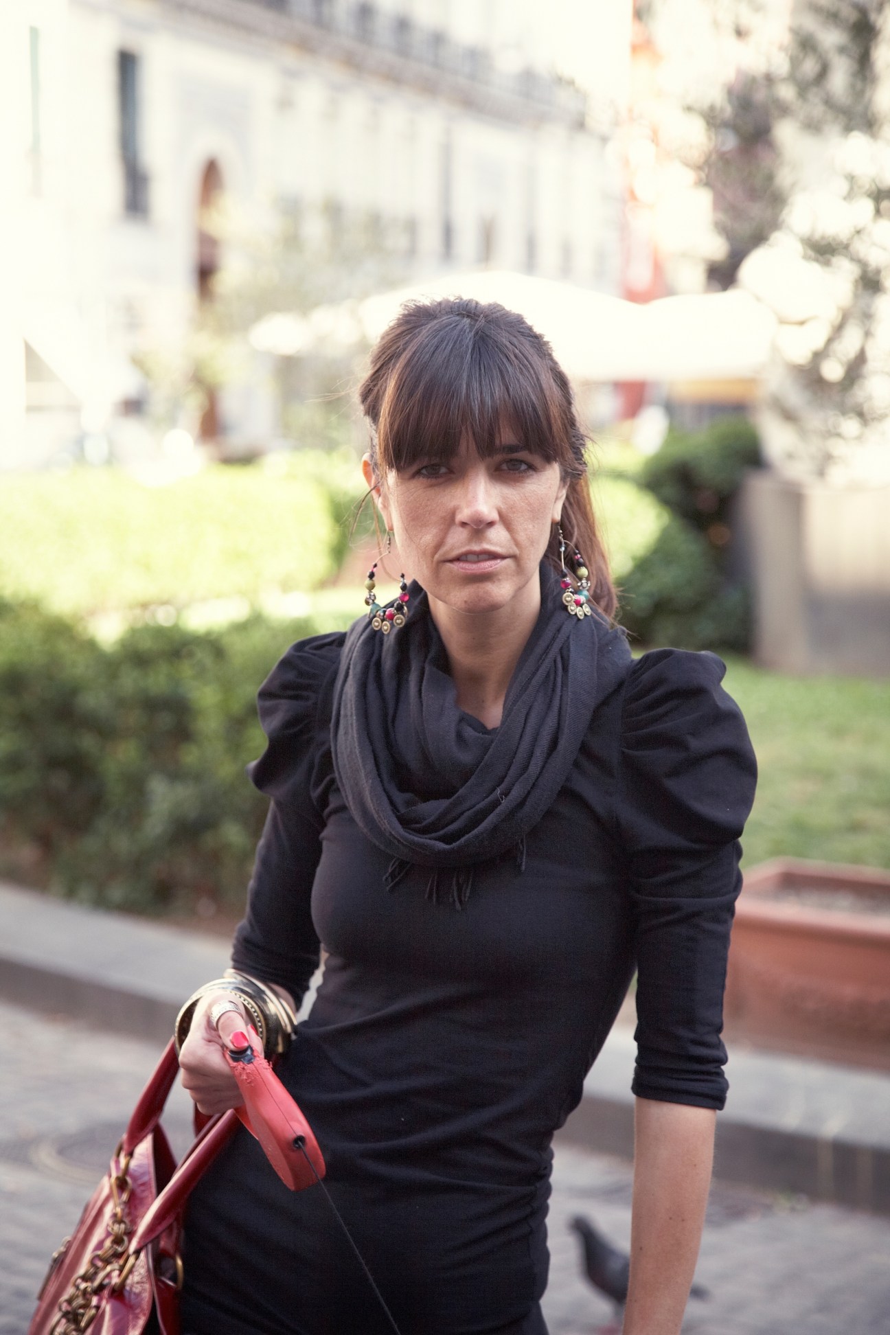 Woman in black clothing and colorful earrings standing in a Naples courtyard with buildings and greenery behind her.