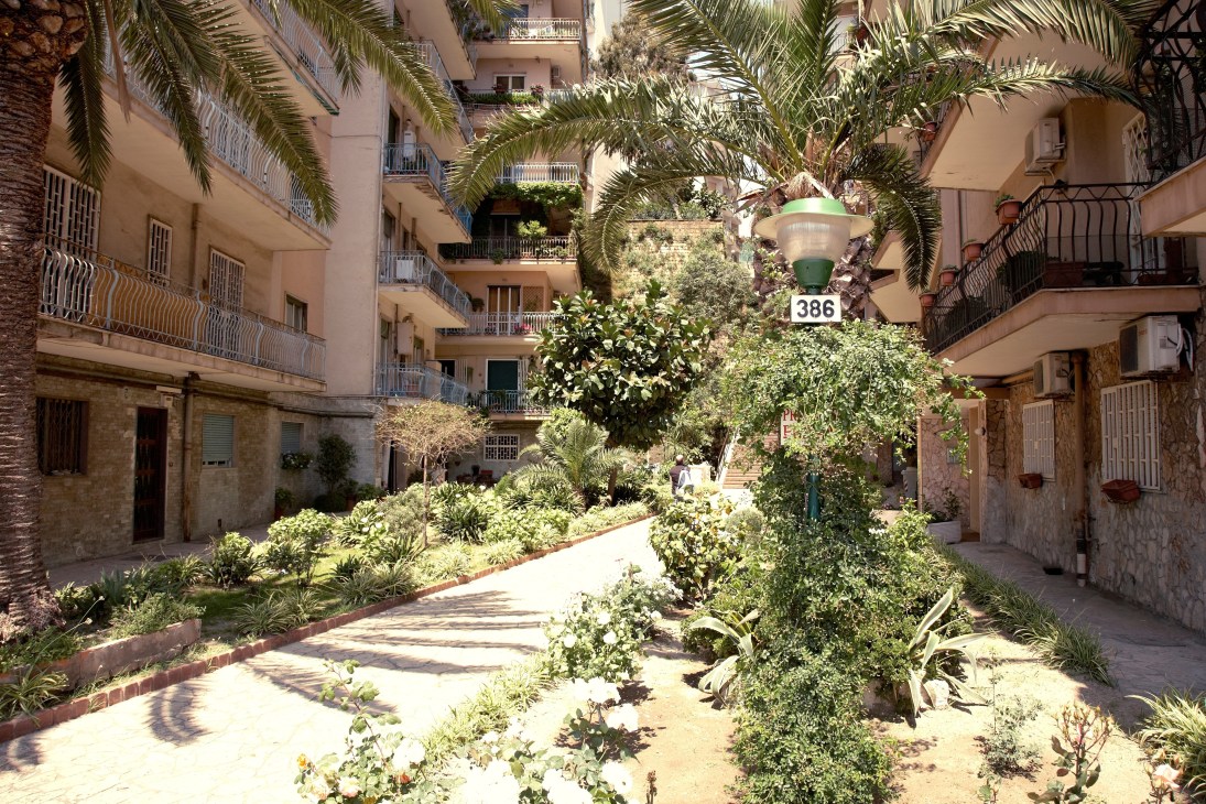 A sunny residential courtyard in Naples surrounded by multi-story apartment buildings with palm trees and lush vegetation.