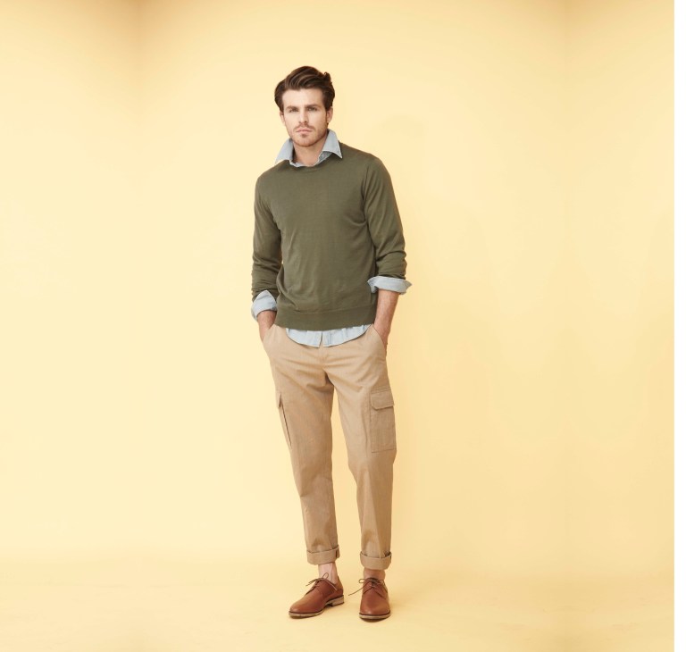 Man wearing olive green sweater, light blue shirt, beige cargo pants and brown shoes against yellow background.
