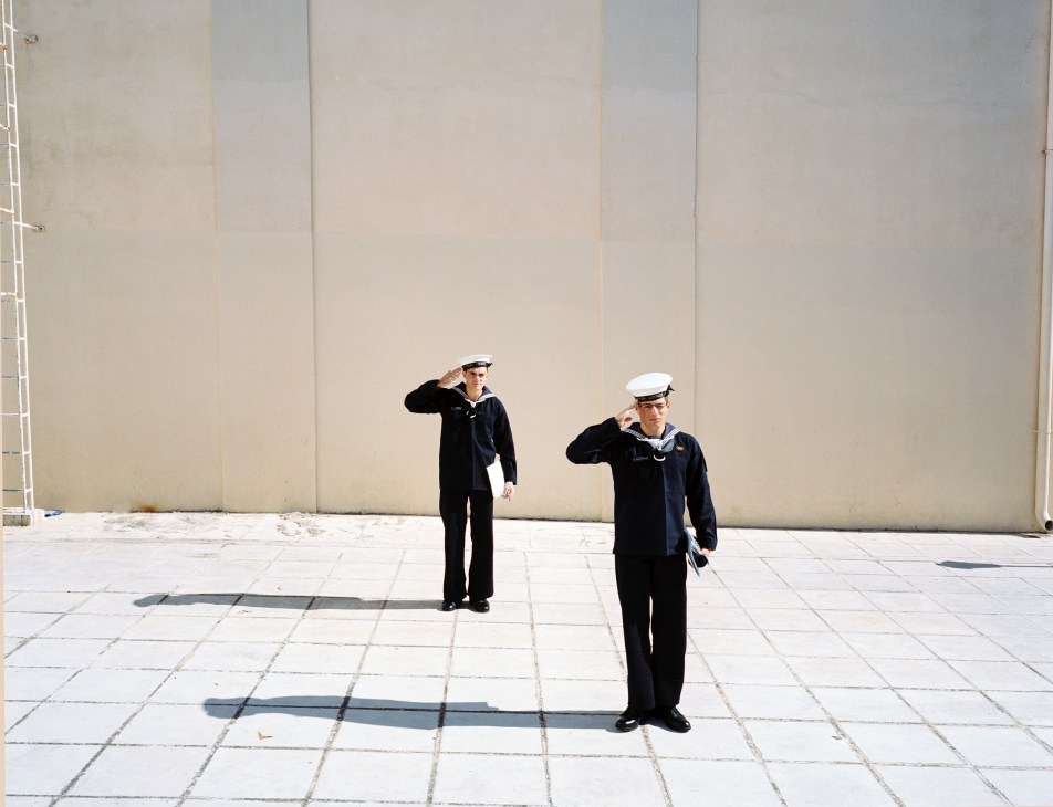 Two naval academy students in dark uniforms and white caps salute on a paved courtyard with beige wall behind them.