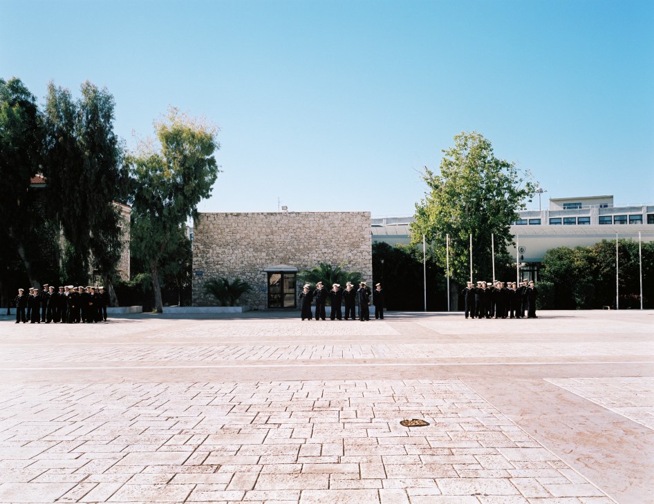 Naval academy students assembled in uniform in a large courtyard surrounded by trees and modern buildings.