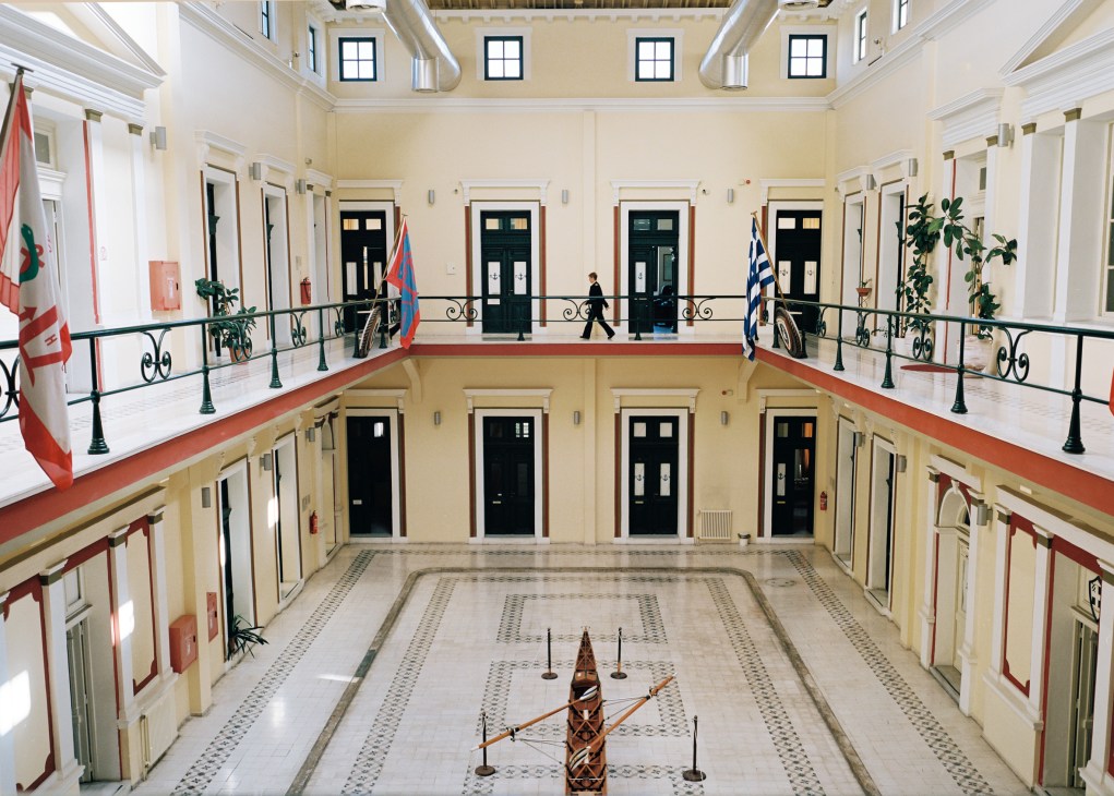Interior courtyard of Piraeus naval academy with cream-colored walls, black doors, red railings, and national flags on upper balconies.