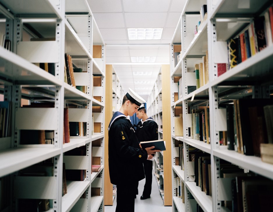 Two naval academy students in uniform browse books in a library with white shelving on both sides.