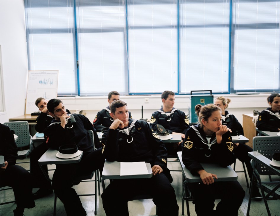 Naval academy students in uniform sit at desks in a classroom with large windows.
