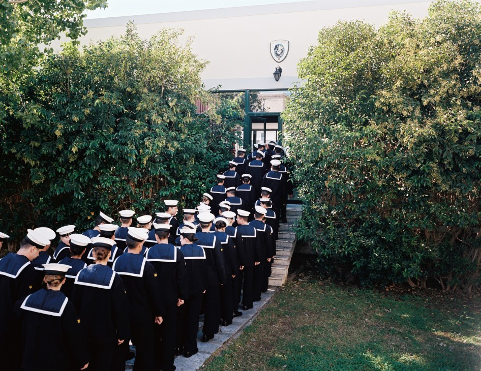 Naval academy students in dark uniforms with white caps stand in an orderly queue between green trees leading to a building entrance.