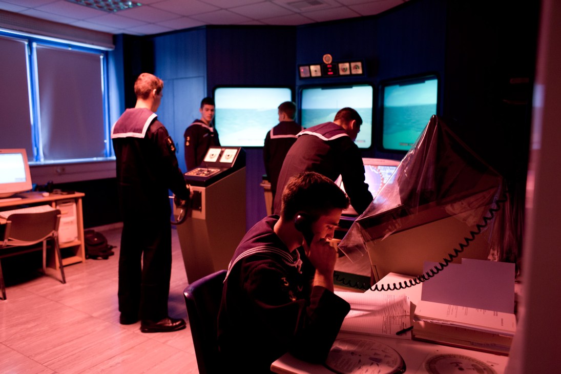 Naval academy students practice navigation in a simulator room with multiple monitors displaying seascapes and control panels.