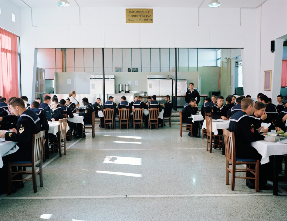 Naval academy students in uniform seated at tables during lunch in a bright refectory with large windows.