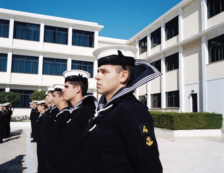 Naval academy students in uniform standing at attention in a courtyard with modern white buildings.