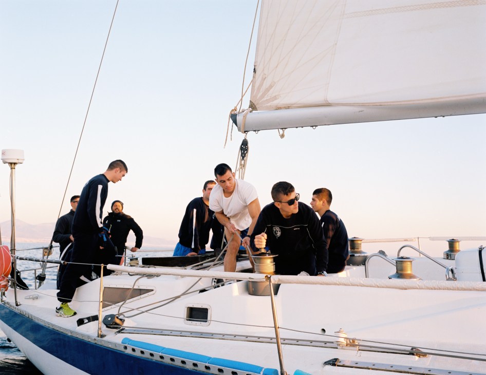Naval academy students practice sailing techniques on a white sailboat with raised sails on calm waters.