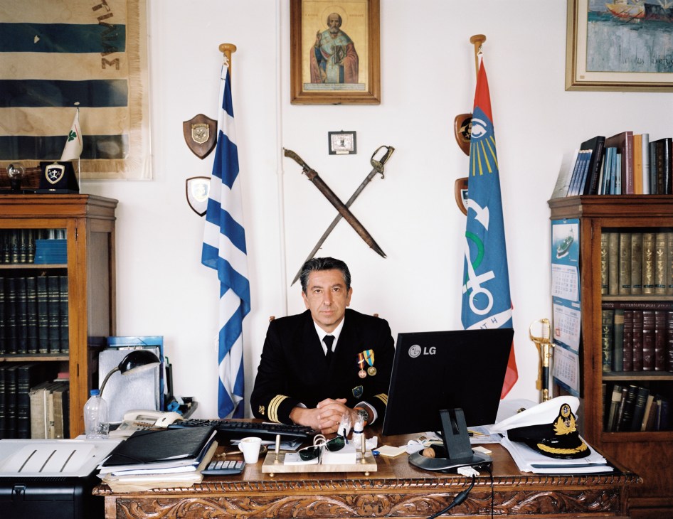 Commanding officer Ioannis Kokios seated at his desk in his office with Greek flags, crossed swords, and maritime artifacts on the wall behind him.
