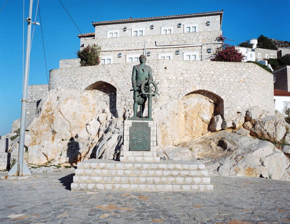 Bronze statue of Andreas Vokos Miaoulis holding a ship's wheel, standing on a stone pedestal against a rocky fortification wall on the Greek island of Hydra.