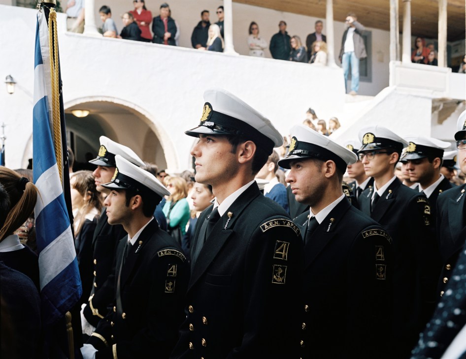 Greek merchant marine cadets in naval uniforms stand at attention during a formal parade with a Greek flag visible.