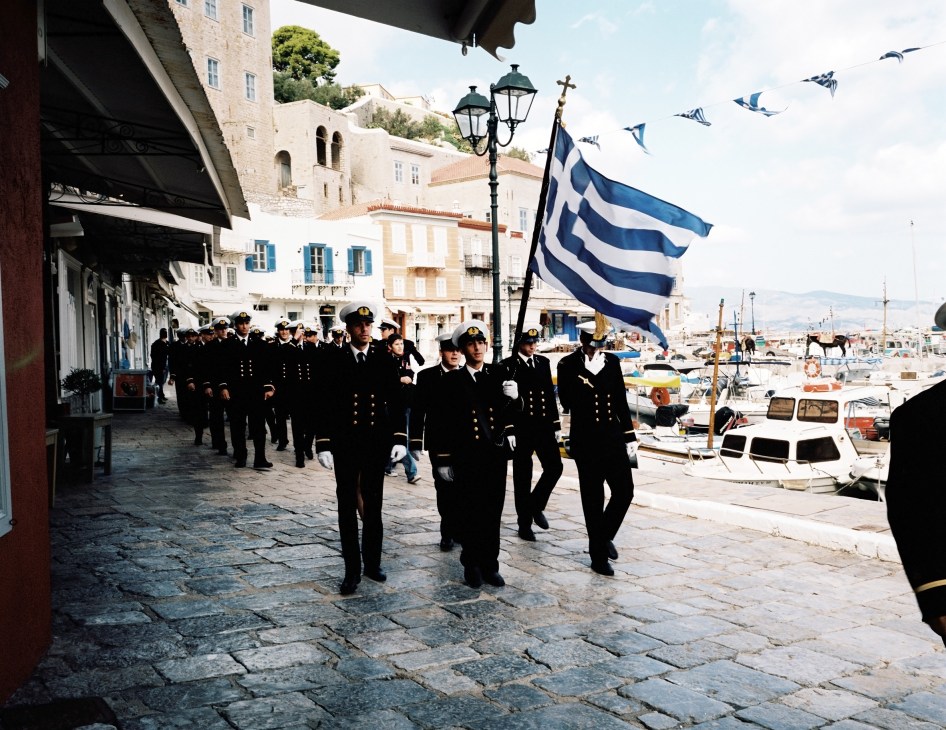 Merchant marine students in dark uniforms march in formation along a waterfront promenade carrying the Greek flag.