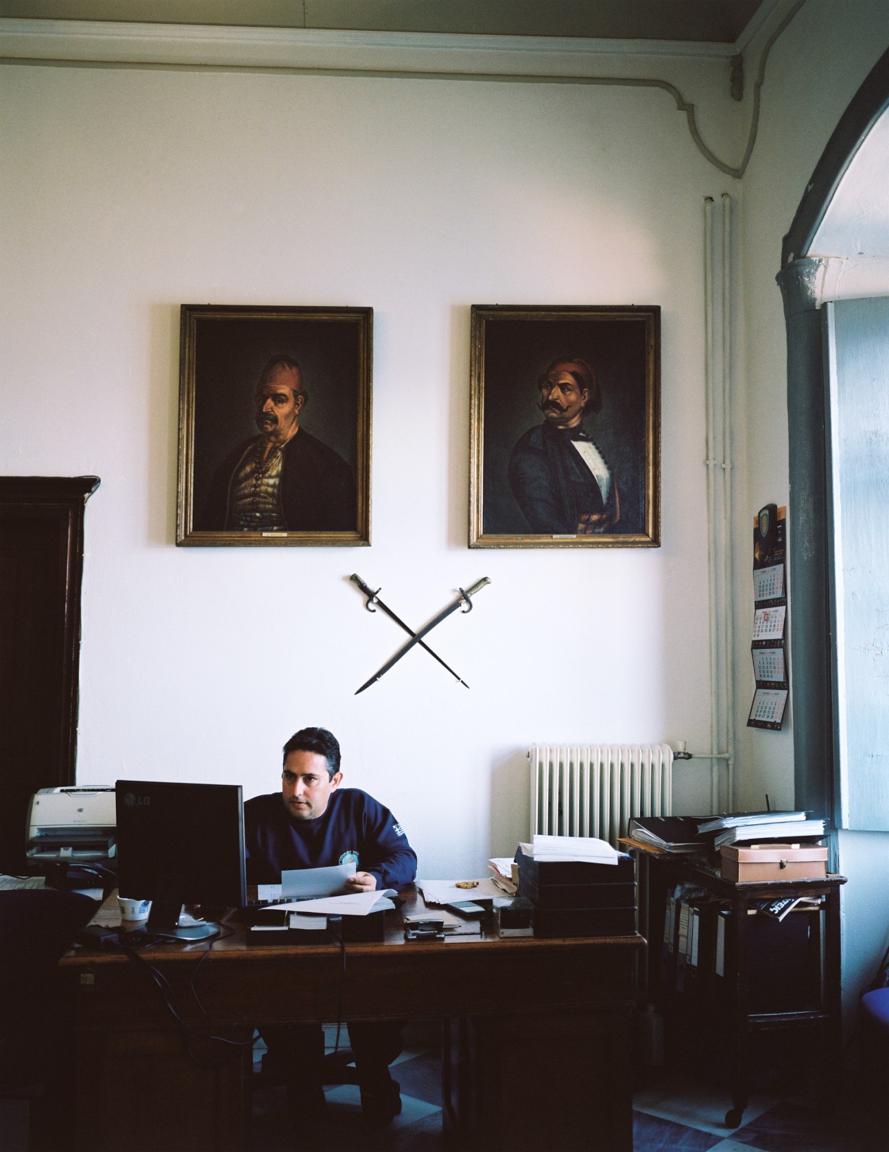 Man working at desk in maritime academy office with historical portraits and crossed swords on wall.
