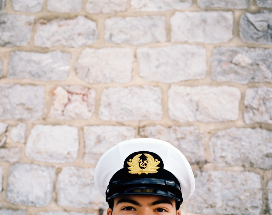 Naval academy student Nikos Papaoikonomou wearing a white cap with gold insignia against a whitewashed stone wall.