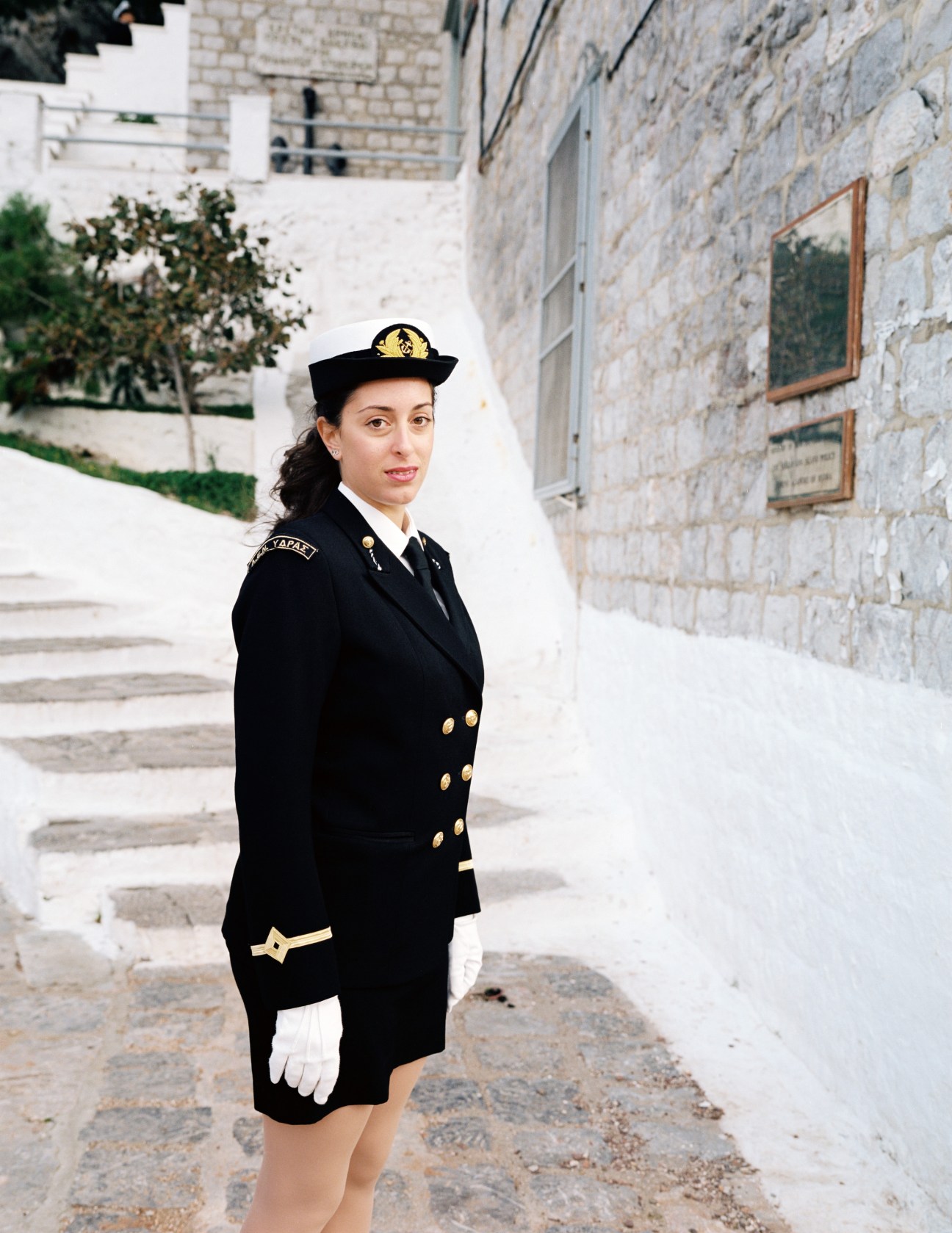 Female naval academy student in black uniform with gold buttons and sailor cap standing in courtyard.
