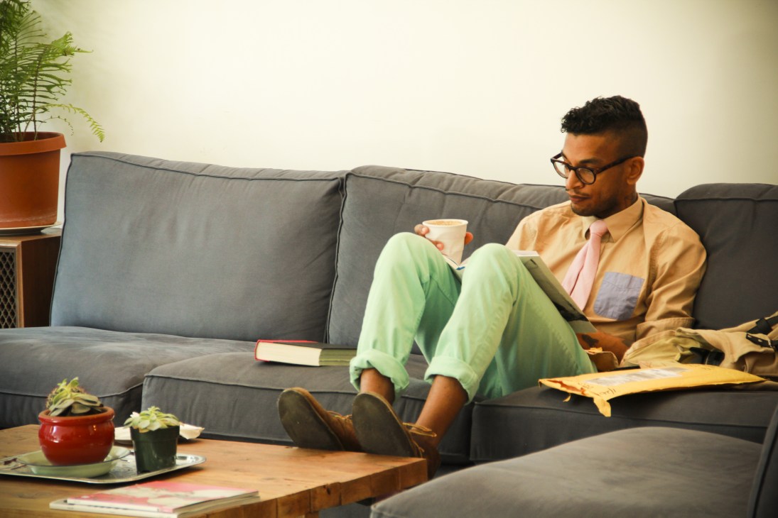 Man sitting on grey sofa reading, holding coffee cup, with books and plants nearby.