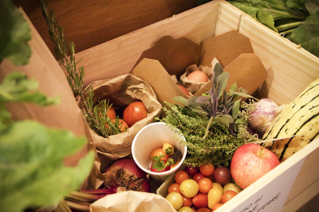 Wooden crate filled with fresh produce including tomatoes, apples, herbs, and vegetables in paper bags.
