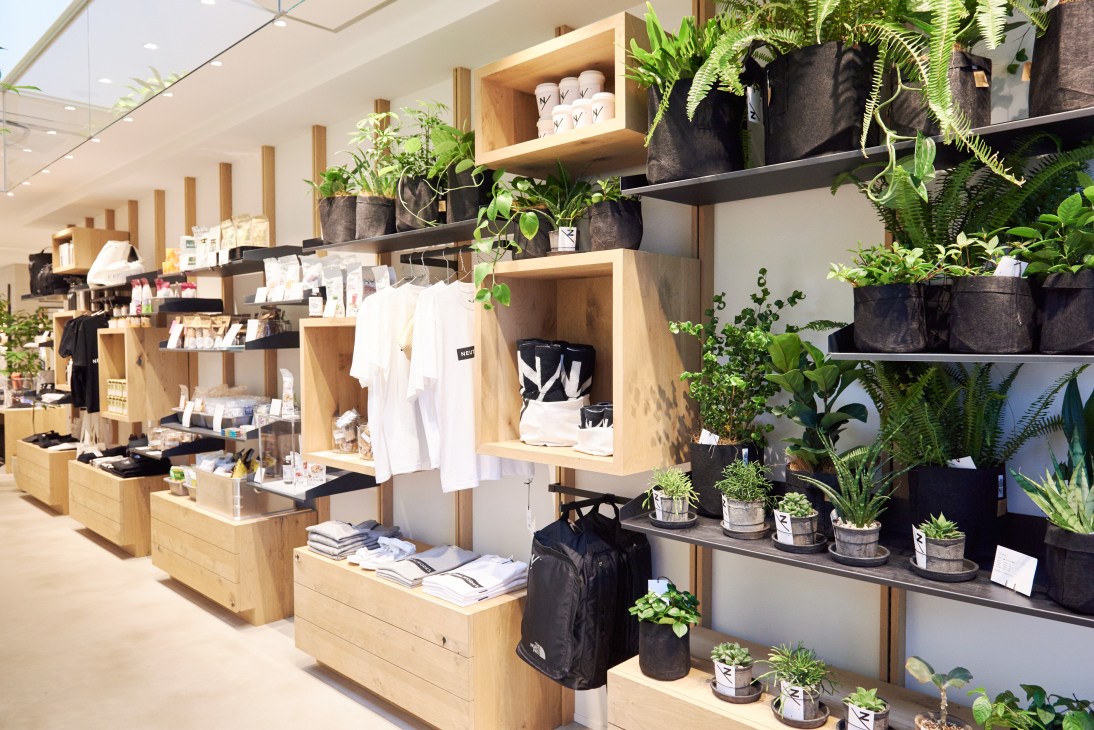 Interior of Neutralworks Tokyo shop with wooden shelving displaying white folded clothing, black backpacks, and potted plants throughout.