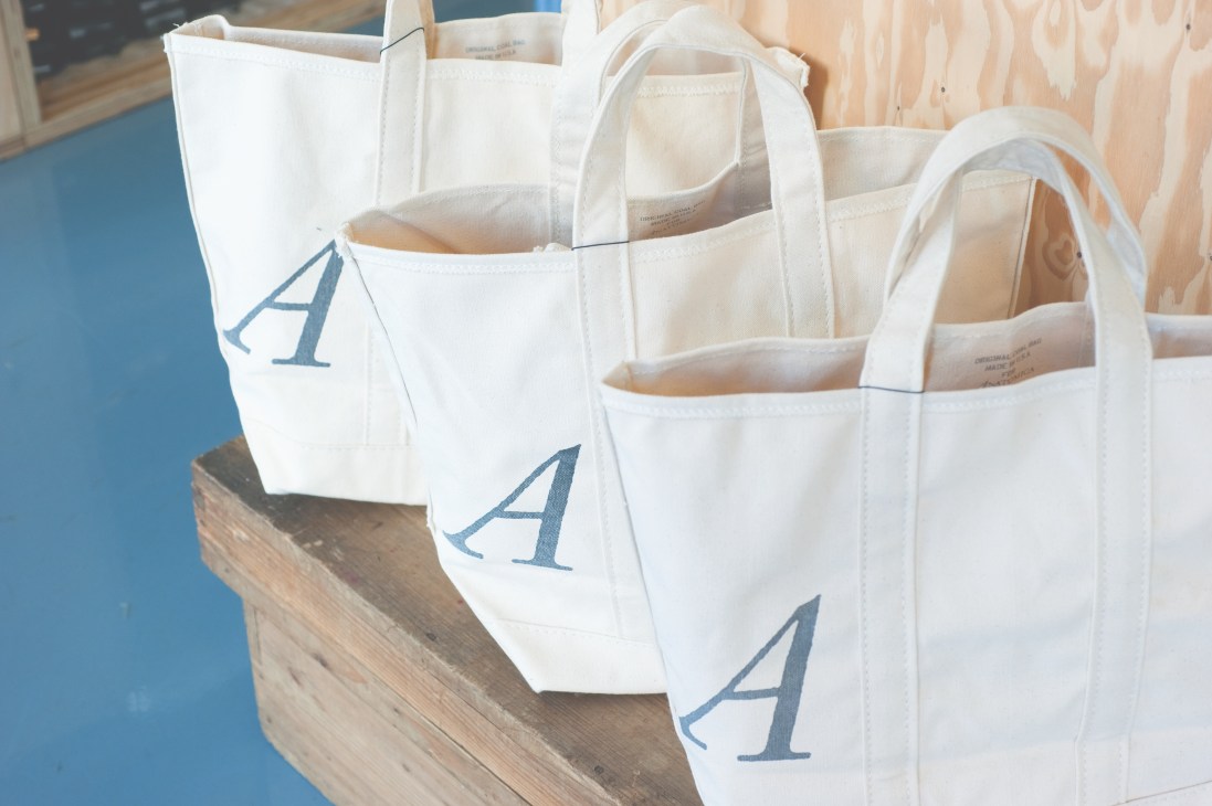 White canvas tote bags with blue 'A' logo displayed on wooden platform.