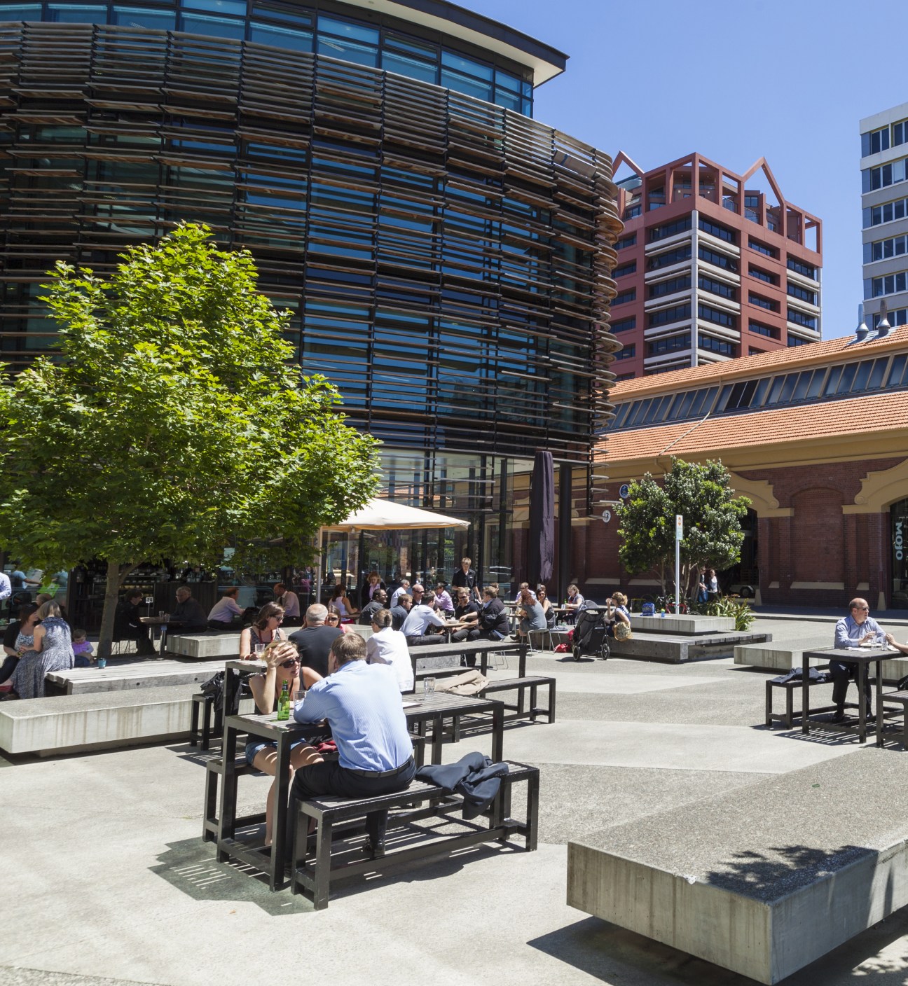 A sunny public plaza in Wellington with people seated at outdoor tables beneath modern buildings.