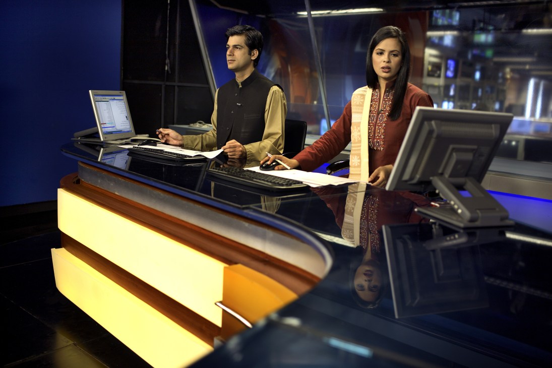 Two news presenters, Khaled and Naija, sit at a modern news desk with computers and monitors in a television studio.