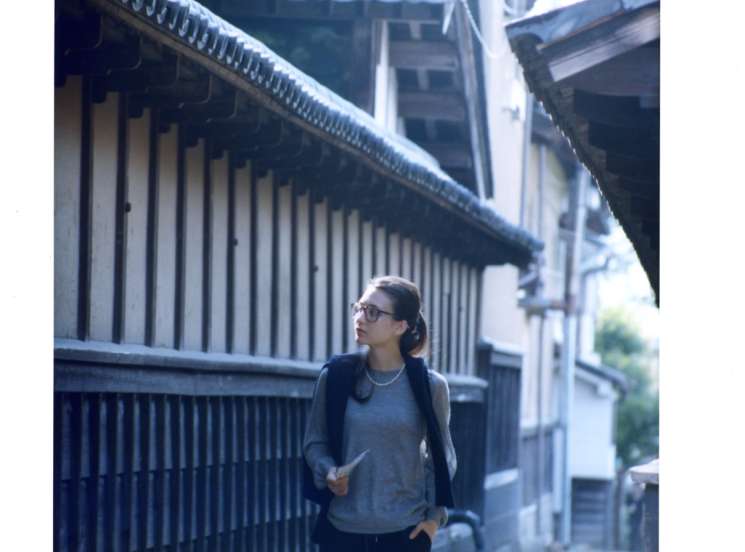 Model wearing Drawer jumper and Bottega Veneta glasses stands in traditional Japanese street with wooden architecture and curved roof tiles.