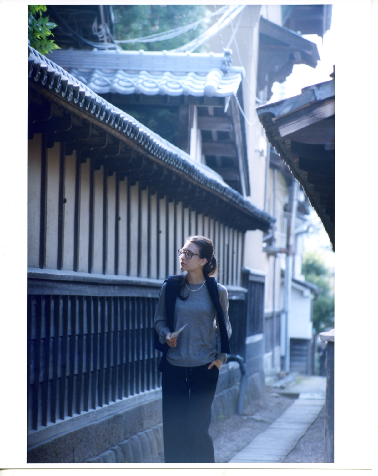Model wearing Drawer jumper and Bottega Veneta glasses stands in traditional Japanese street with wooden architecture and curved roof tiles.