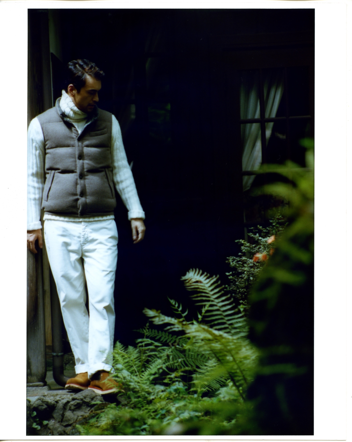 Man wearing Brunello Cucinelli vest and Boss Selection jumper standing among ferns in a shaded garden setting.