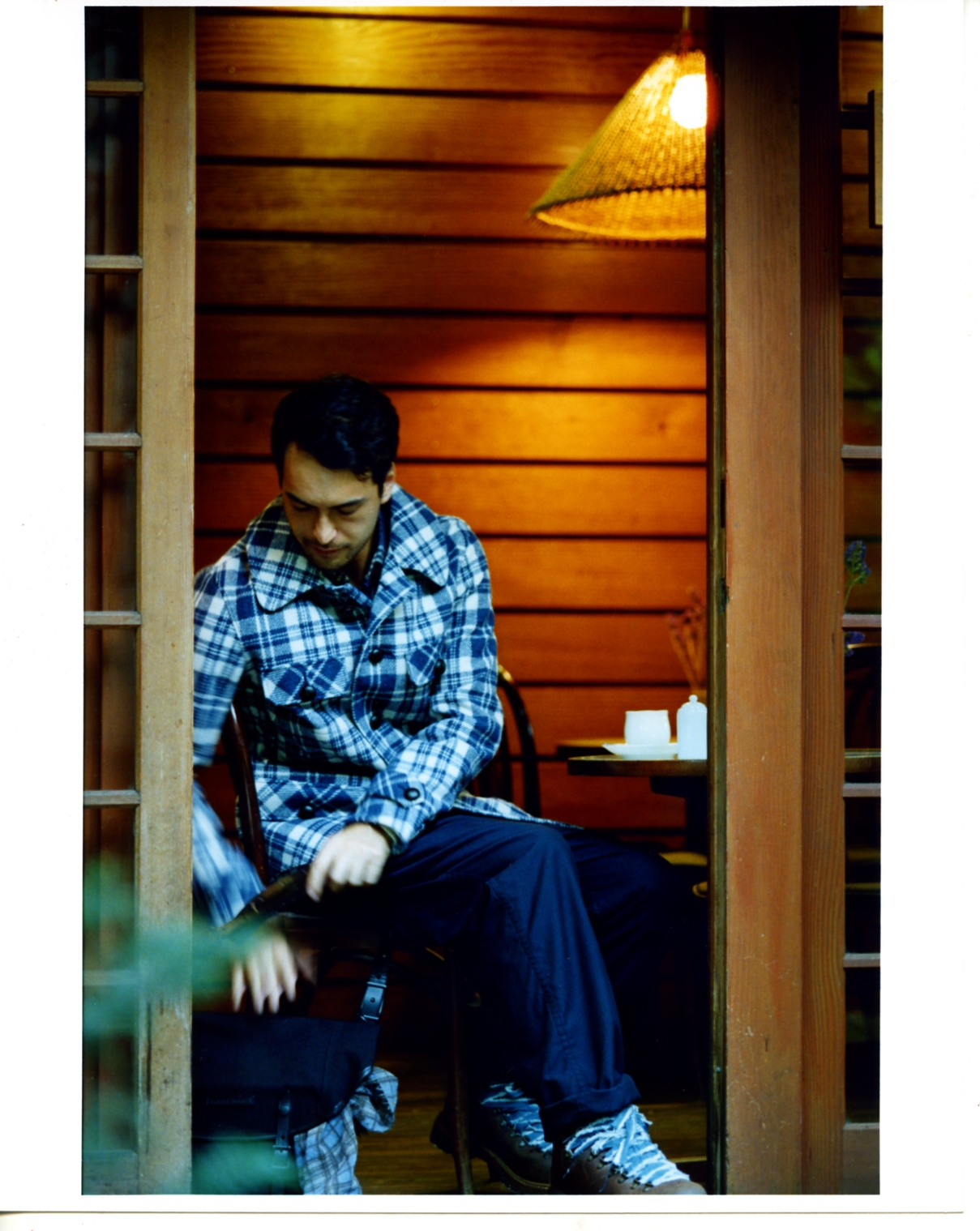 Model wearing blue and white plaid shirt and dark trousers seated in wooden alcove with warm lighting.