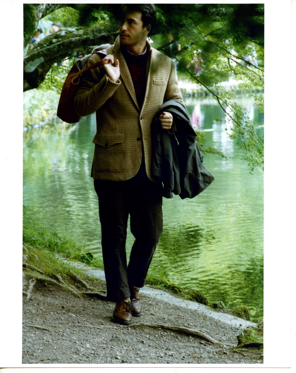 Man in beige blazer carrying black coat walks along pond path lined with green foliage.