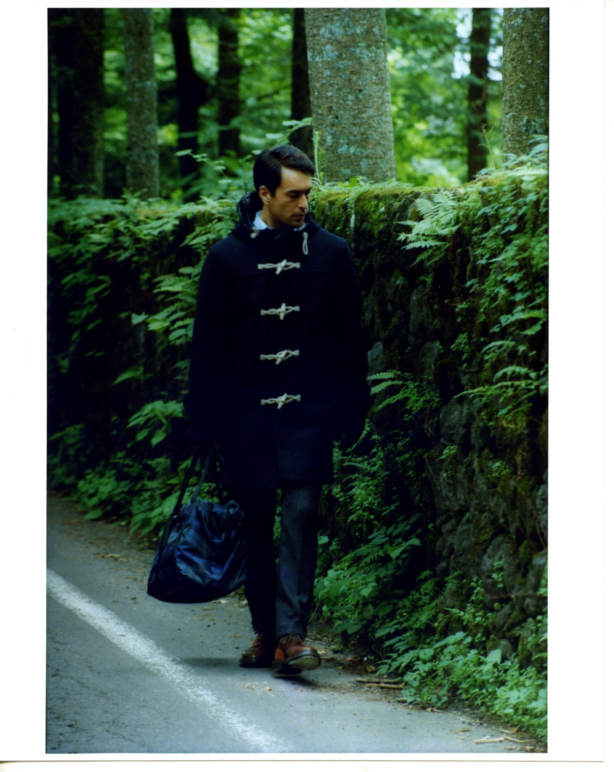 Man wearing a black Nanamica coat with toggle buttons walks on a forest path lined with moss-covered rocks and ferns.