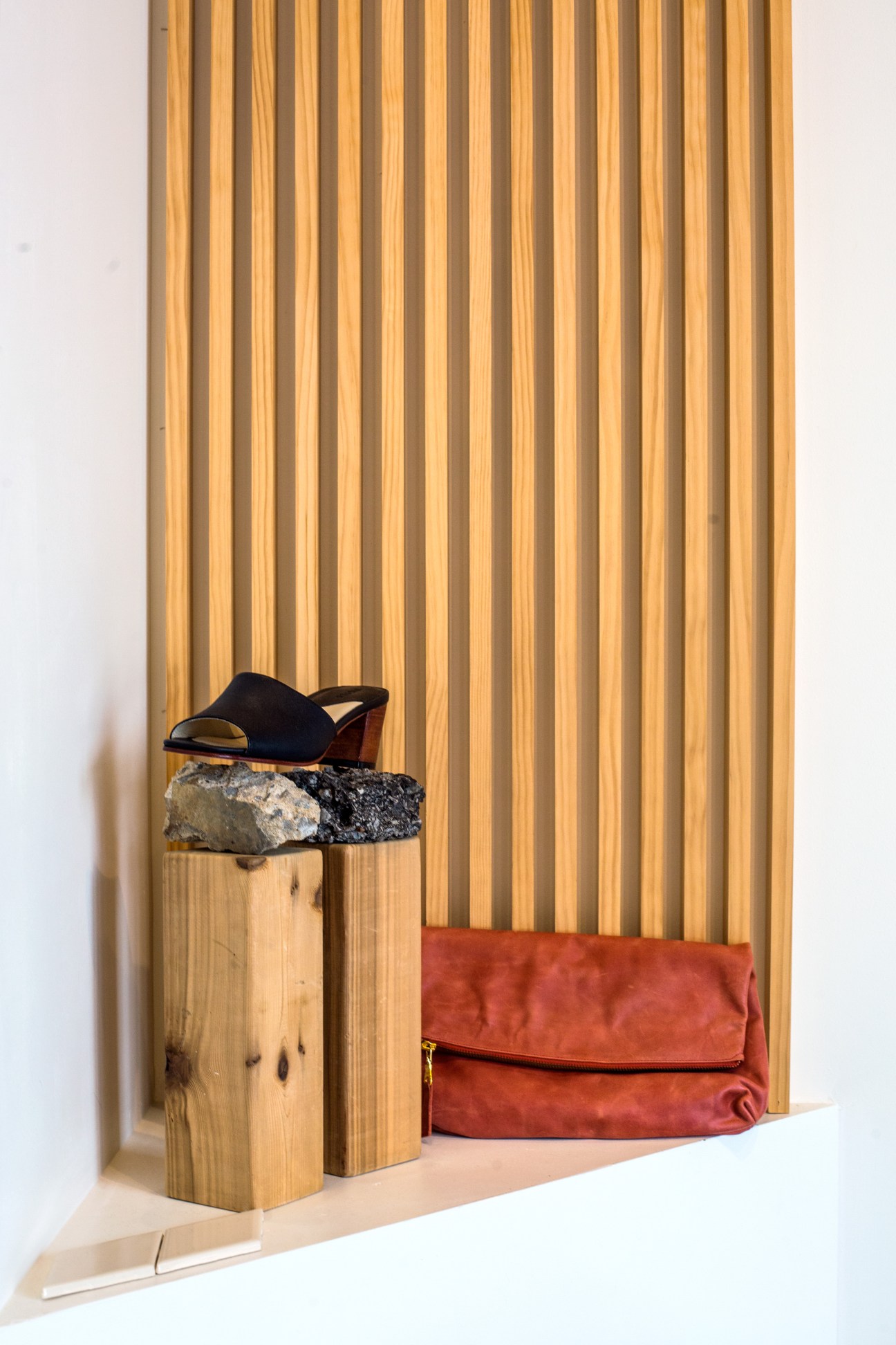 Black suede mule shoe and red leather clutch displayed on wooden blocks against corrugated metal backdrop.