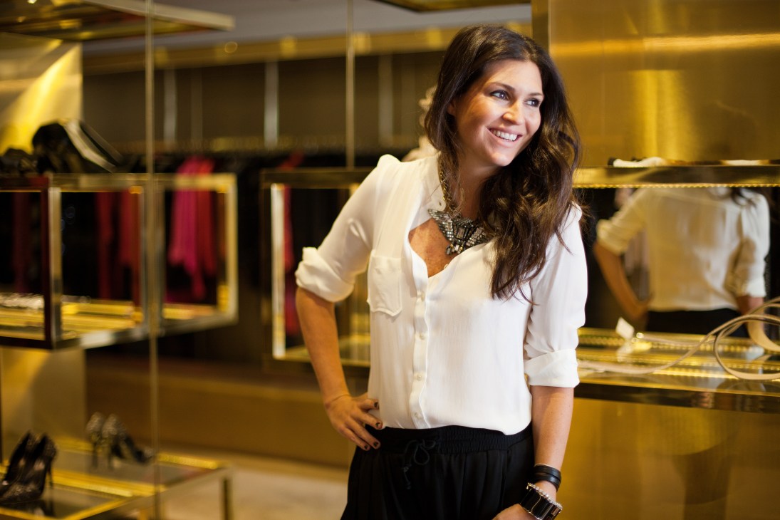 Natalie Klein, founder of NK boutique, smiling in her luxury retail store with clothing displays and gold fixtures visible in the background.