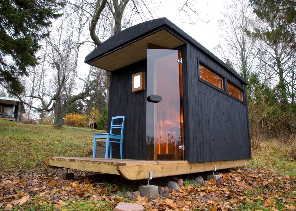 Portable black pine sauna with large windows and wooden deck, designed by Denizen, positioned in a forest setting.