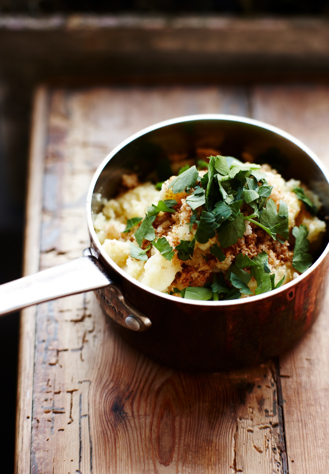 Creamy potato dish with crispy breadcrumb topping and fresh parsley in a white-rimmed cooking pot on wooden surface.