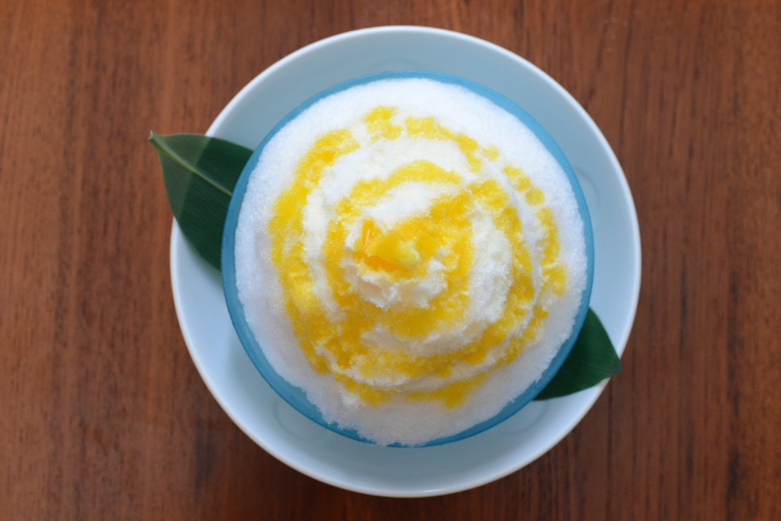 Shaved ice with condensed milk and yuzu sorbet in a blue-rimmed bowl, garnished with a green leaf.