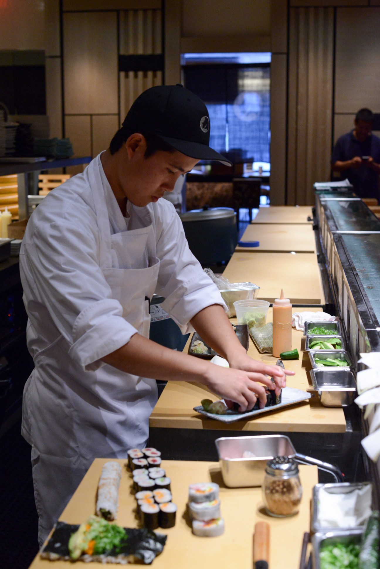 Chef in white apron and black cap prepares a dish at the sushi bar counter.